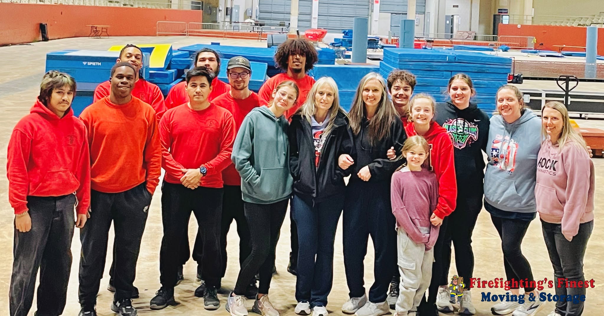 The Firefighting's Finest Moving & Storage team in red shirts posing with local community members at a large gymnasium in Plano, TX.