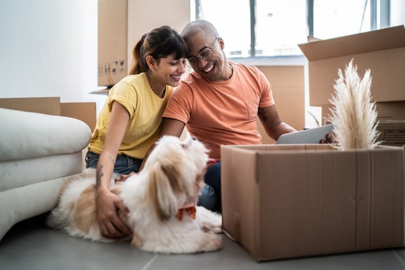 Mixed race couple sitting on floor, surrounded by moving boxes and their dog